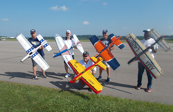 Five people holding model airplanes on a tarmac, smiling on a sunny day.